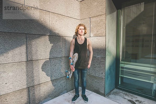 Portrait eines jungen männlichen Skateboardfahrers  der in einer Ecke steht und Skateboard hält.