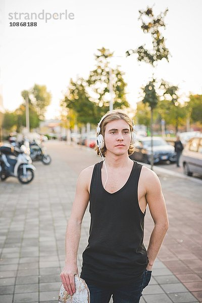 Portrait eines jungen männlichen Skateboardfahrers  der Kopfhörermusik auf dem Bürgersteig hört.