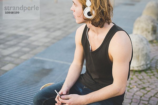 Junger Mann sitzt auf dem Bürgersteig und hört Kopfhörermusik.