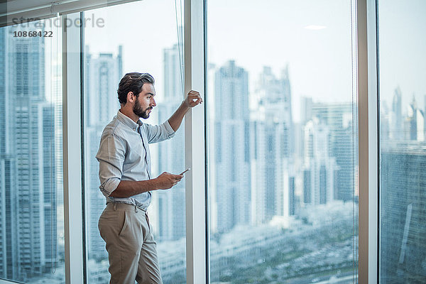 Geschäftsmann mit Smartphone  der durch ein Fenster mit Blick auf einen Wolkenkratzer starrt  Dubai  Vereinigte Arabische Emirate