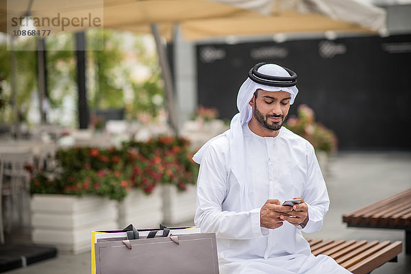 Männlicher Einkäufer in traditioneller Kleidung aus dem Nahen Osten sitzt auf Bank und liest Smartphone-Text  Dubai  Vereinigte Arabische Emirate