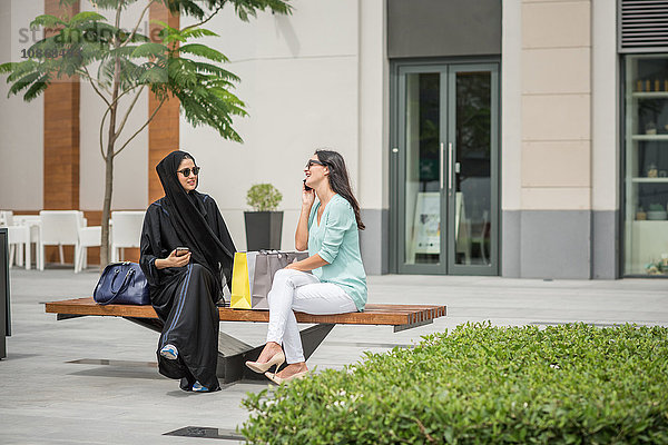 Junge Frau aus dem Nahen Osten in traditioneller Kleidung mit Freundin auf Bank sitzend  Dubai  Vereinigte Arabische Emirate