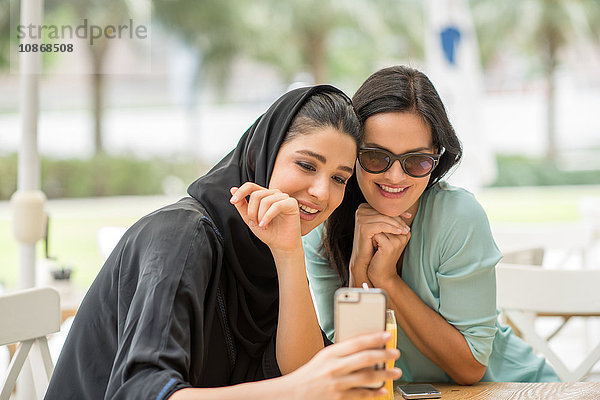 Junge Frau aus dem Nahen Osten in traditioneller Kleidung liest mit einer Freundin im Café einen Text auf einem Smartphone  Dubai  Vereinigte Arabische Emirate