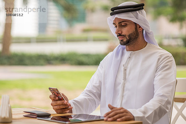 Mann aus dem Nahen Osten in traditioneller Kleidung liest Smartphone-Text im Café  Dubai  Vereinigte Arabische Emirate