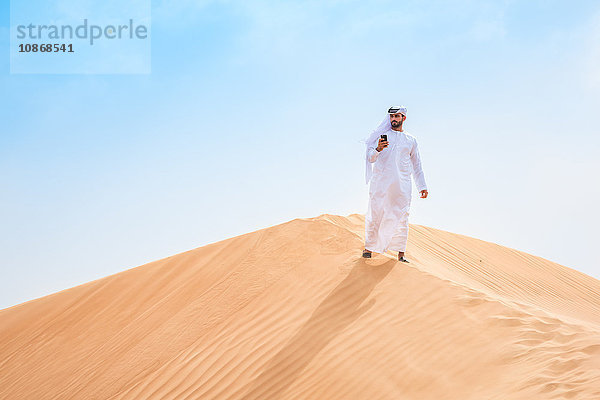 Mann aus dem Nahen Osten trägt traditionelle Kleidung mit Smartphone auf Wüstendüne  Dubai  Vereinigte Arabische Emirate