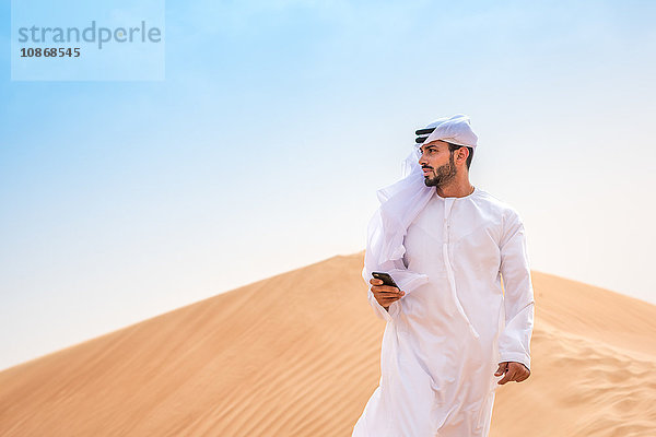 Mann aus dem Nahen Osten trägt traditionelle Kleidung mit Smartphone auf Wüstendüne  Dubai  Vereinigte Arabische Emirate