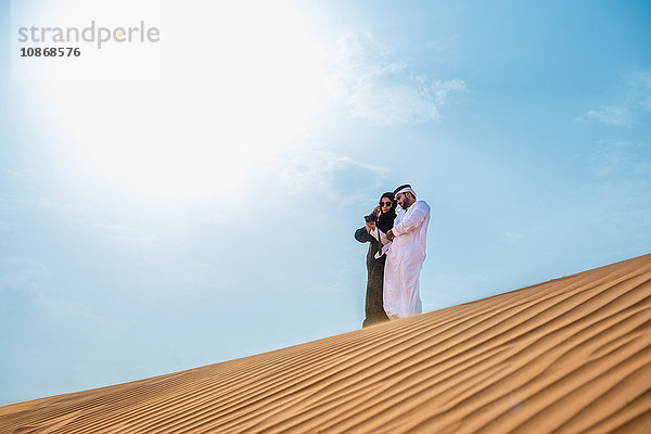 Paar aus dem Nahen Osten in traditioneller Kleidung mit Smartphone-Selfie auf Wüstendüne  Dubai  Vereinigte Arabische Emirate