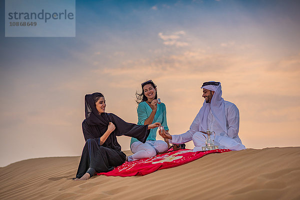 Einheimisches Paar in traditioneller Kleidung picknickt mit Touristin auf Wüstendüne  Dubai  Vereinigte Arabische Emirate
