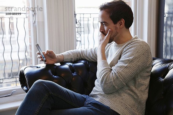 Mittelgroßer erwachsener Mann sitzt auf dem Sofa und schaut auf ein Smartphone