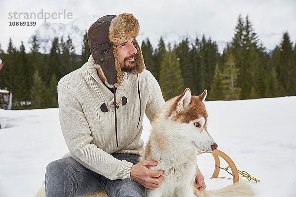 Junger Mann mit Trapperhut streichelt Husky im Schnee  Elmau  Bayern  Deutschland