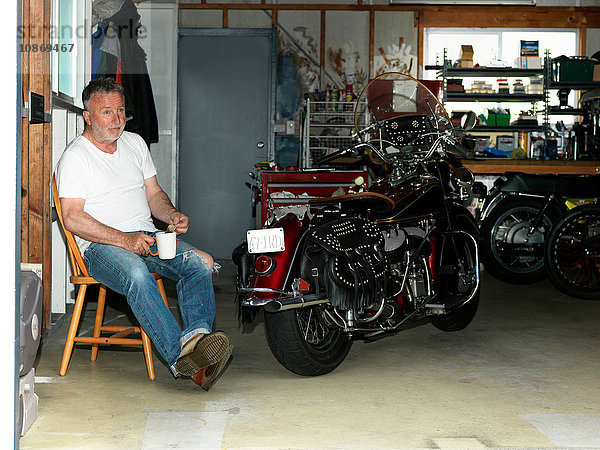 Älterer Mann sitzt mit einem Kaffee in der Garage mit Motorrad