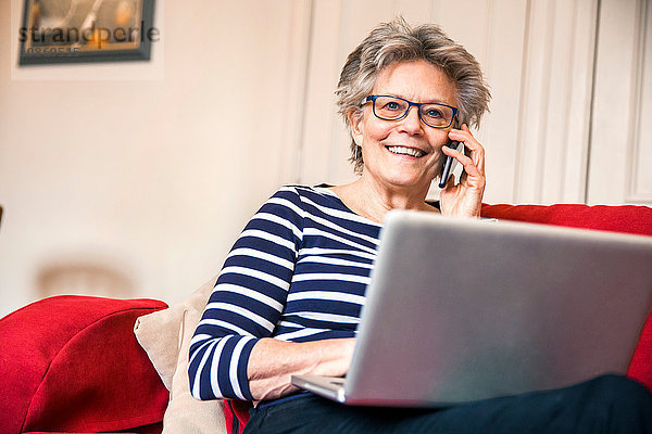 Ältere Frau auf Wohnzimmer-Sofa  die sich mit einem Smartphone unter Verwendung eines Laptops unterhält