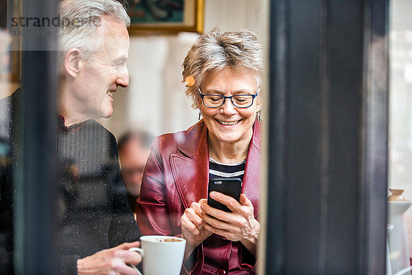 Älteres Ehepaar sitzt am Fensterplatz eines Cafés  trinkt Kaffee und simst per Smartphone
