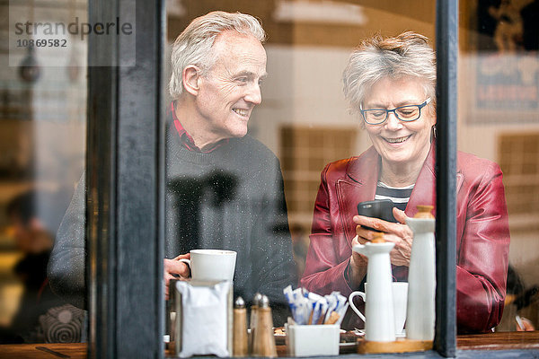 Älteres Ehepaar am Fensterplatz eines Cafés  trinkt Kaffee und schreibt SMS auf Smartphone