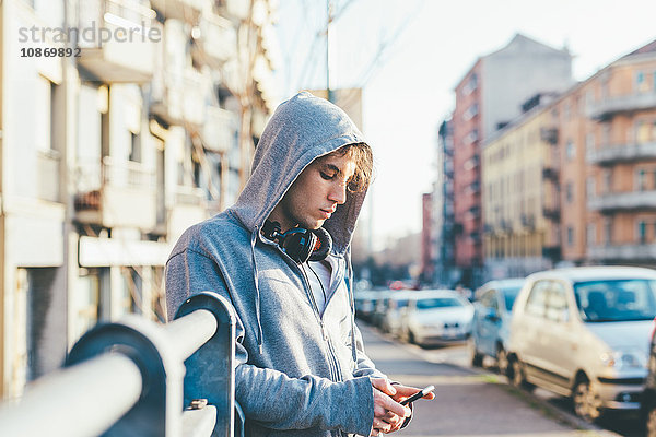 Mann in Stadtgebiet mit Kapuzenoberteil und Kopfhörern schaut auf Smartphone