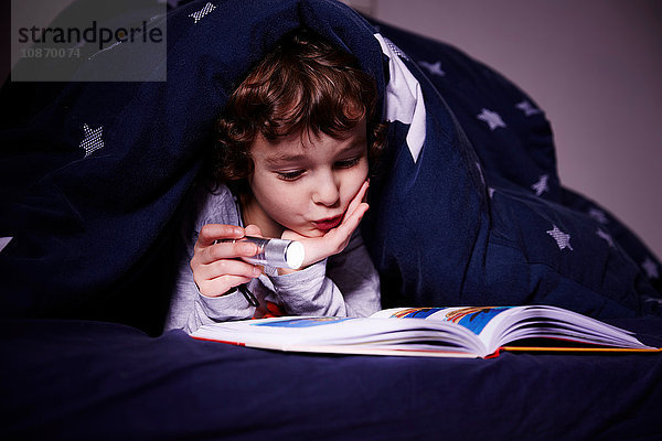 Junge unter der Bettdecke liest Buch bei Fackelschein