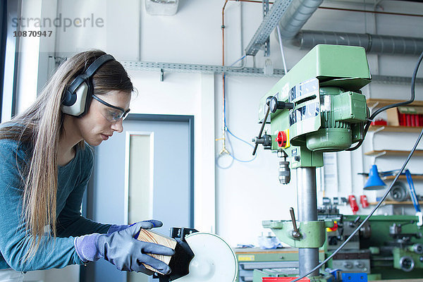 Seitenansicht einer Frau in der Werkstatt  die Gehörschützer trägt und eine Bandschleifmaschine bedient