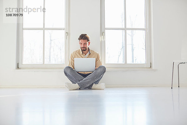 Junger Mann sitzt auf dem Boden und stöbert am Laptop in einem leeren neuen Haus