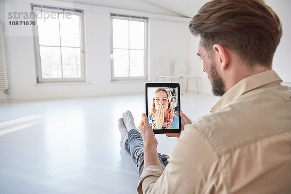 Freundin auf digitalem Tablet-Videoanruf  die einem auf dem Boden sitzenden jungen Mann einen Kuss gibt