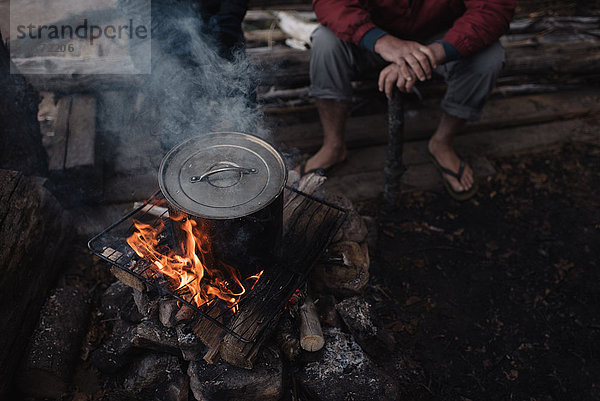 Älterer Mann sitzt auf einem Holzklotz und schaut dem Kochtopf am Lagerfeuer zu  niedriger Abschnitt