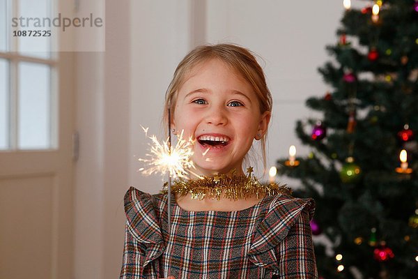 Mädchen vor dem Weihnachtsbaum mit Wunderkerze in der Hand und lächelndem Blick in die Kamera