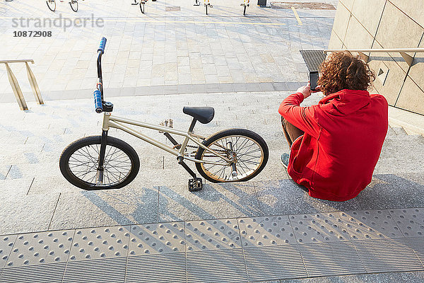 Rückansicht eines auf Stufen sitzenden Mannes mit BMX und Smartphone