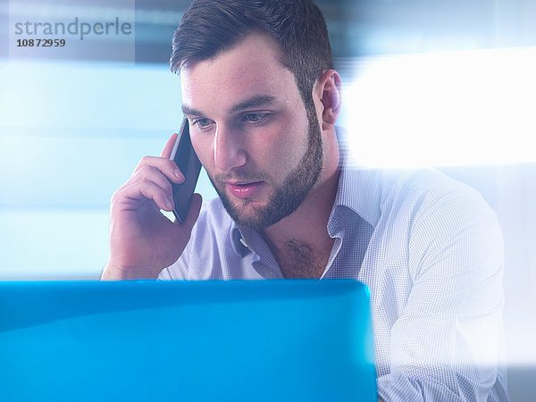 Junger Geschäftsmann spricht auf seinem Smartphone  während er im Büro am Laptop arbeitet