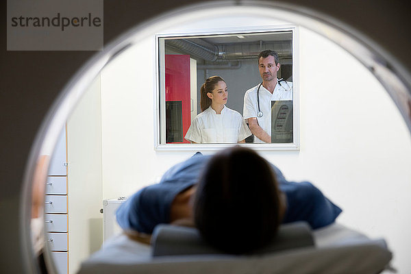 Arzt und Krankenschwester beobachten weibliche CT-Patientin vom Fenster des Kontrollraums