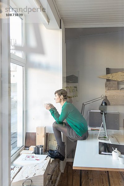 Blick durch Glas  Seitenansicht in voller Länge einer reifen Frau  die auf einem Schreibtisch sitzt und aus dem Fenster schaut