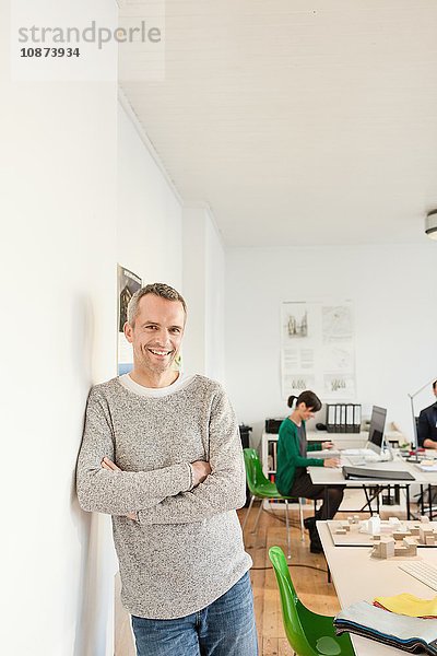 Reifer Mann im Büro lehnt an Wand  die Arme verschränkt und lächelnd in die Kamera blickend