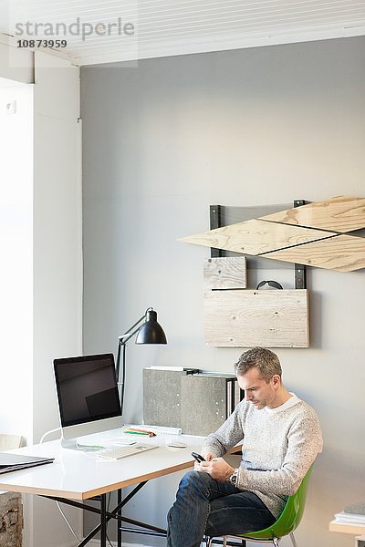 Reifer Mann sitzt im Büro am Schreibtisch und schaut mit seinem Smartphone nach unten