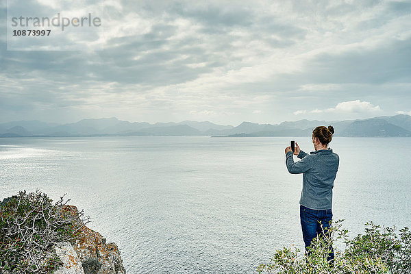 Junger Mann steht auf Klippe und fotografiert mit Smartphone  Alcudia  Mallorca  Spanien