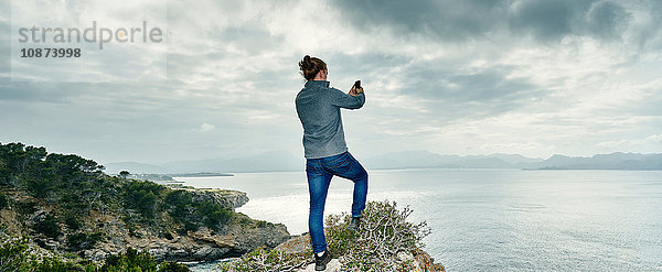 Panoramabild eines jungen Mannes  der auf einer Klippe steht und mit einem Smartphone fotografiert  Alcudia  Mallorca  Spanien