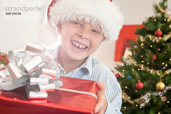Junge mit Weihnachtsmannhut hält Weihnachtsgeschenk und schaut lächelnd in die Kamera
