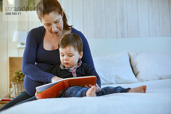 Schwangere Mutter und kleiner Junge sitzen zusammen auf dem Bett und lesen Buch