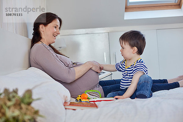 Seitenansicht eines kleinen Jungen auf dem Bett  der den schwangeren Bauch der Mutter berührt