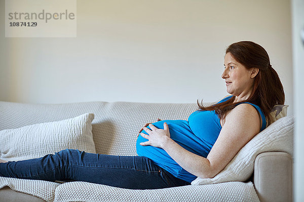 Seitenansicht einer schwangeren Frau  die den Bauch hält  sich auf dem Sofa entspannt und lächelnd wegschaut