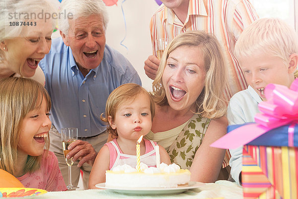 Kleines Mädchen umgeben von Familie bläst Kerze auf Geburtstagskuchen aus