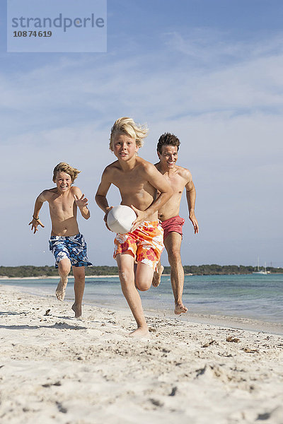 Junge rennt mit Rugbyball  der von Bruder und Vater am Strand verfolgt wird  Mallorca  Spanien