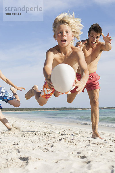 Junge springt in der Luft mit einem Rugbyball  der von Bruder und Vater am Strand verfolgt wird  Mallorca  Spanien