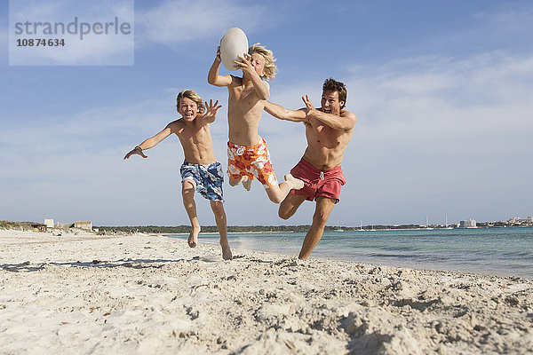 Junge springt mit Rugbyball  der von Bruder und Vater am Strand verfolgt wird  Mallorca  Spanien