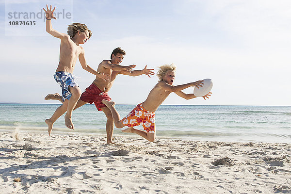 Junge und Vater jagen Bruder mit Rugbyball am Strand hinterher  Mallorca  Spanien