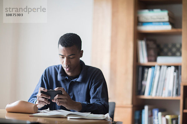 Junger männlicher Gymnasiast sitzt am Schreibtisch und liest Smartphone-Texte