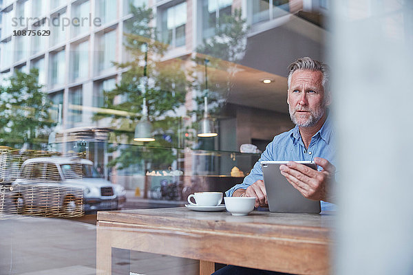 Reifer Mann sitzt im Café  benutzt digitales Tablett  Straße spiegelt sich im Fenster