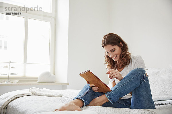 Schöne junge Frau sitzt auf Bett und benutzt digitales Tablett