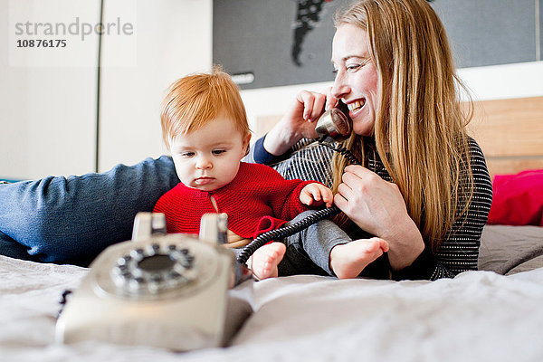 Mittelgroße erwachsene Frau und kleine Tochter im Bett sprechen am Festnetztelefon