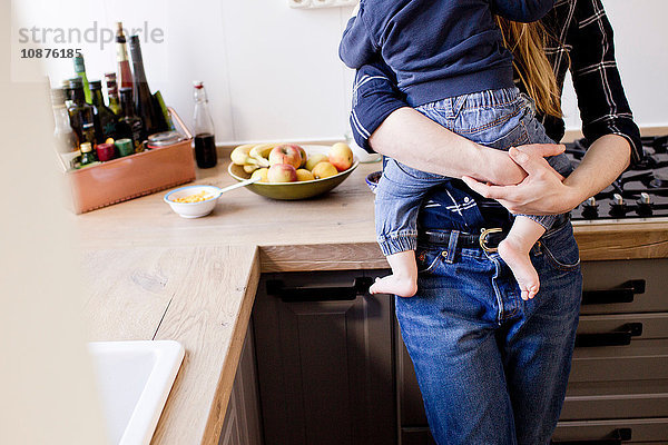 Schnappschuss einer mittelgroßen erwachsenen Frau  die einen kleinen Sohn in der Küche trägt