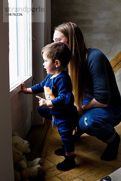 Mittelgroße erwachsene Frau und kleiner Sohn schauen durch das Wohnzimmerfenster