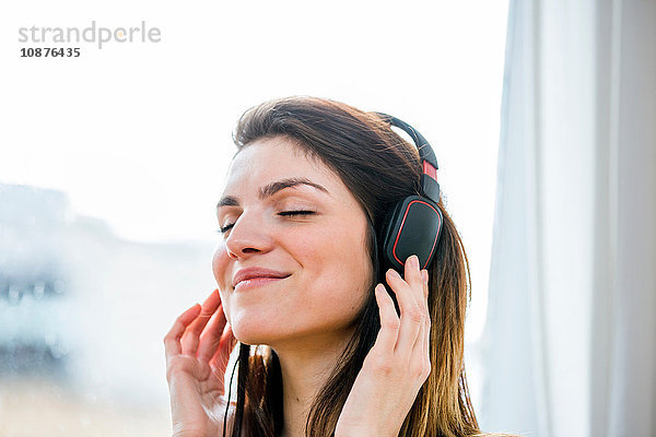 Schöne junge Frau hört Kopfhörermusik vor dem Wohnungsfenster
