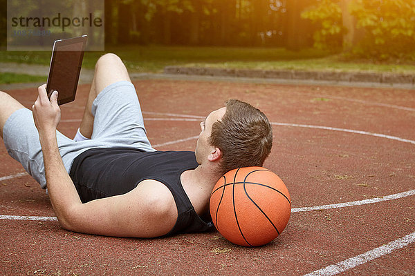 Junger männlicher Basketballspieler liegt auf dem Platz und stöbert auf einem digitalen Tablet